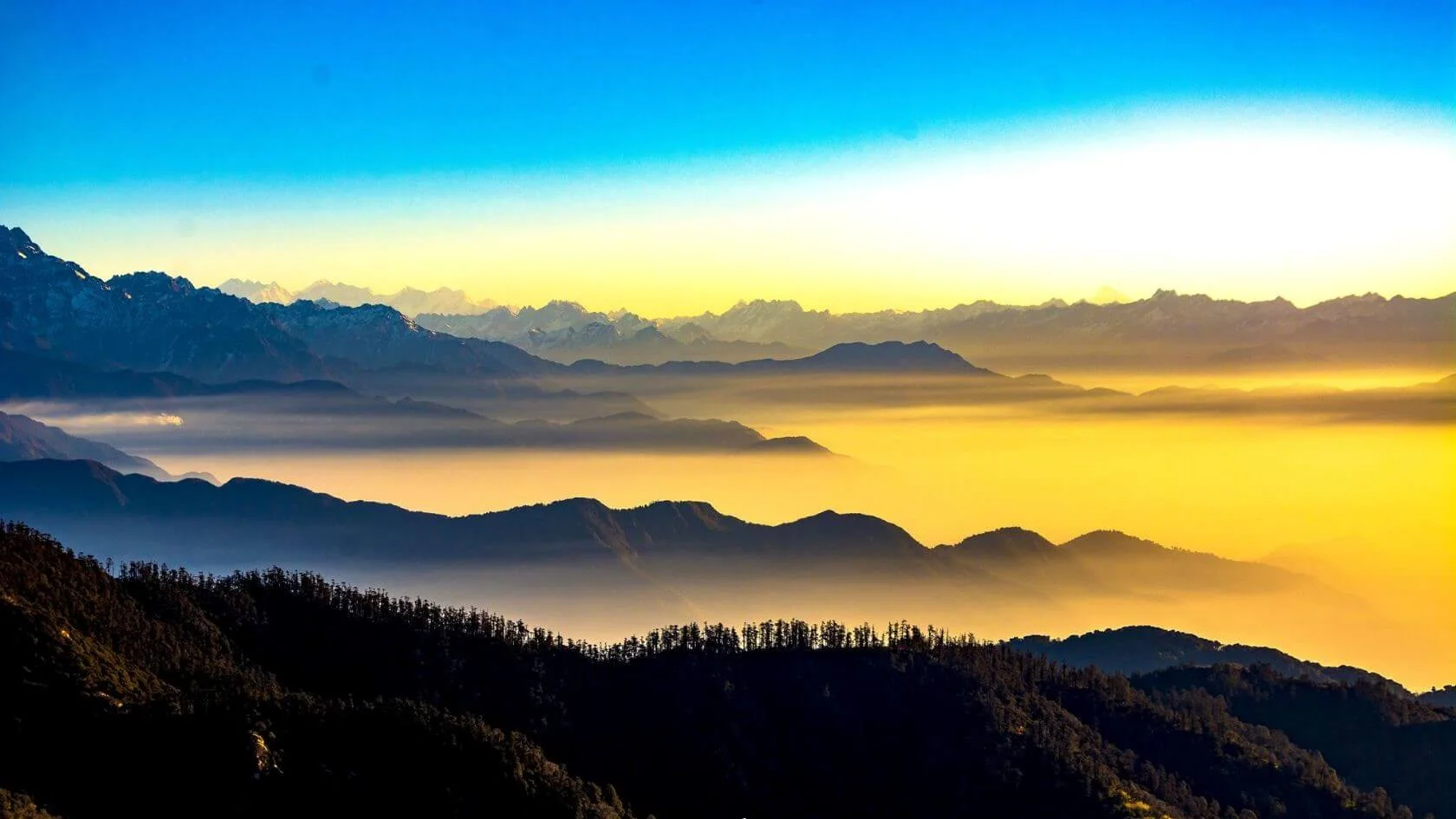 A foggy valley with mountains in the background during the yellow and orange sunset of Darjeeling. The mountains are shrouded in mist showing one of the best times to visit Darjeeling.