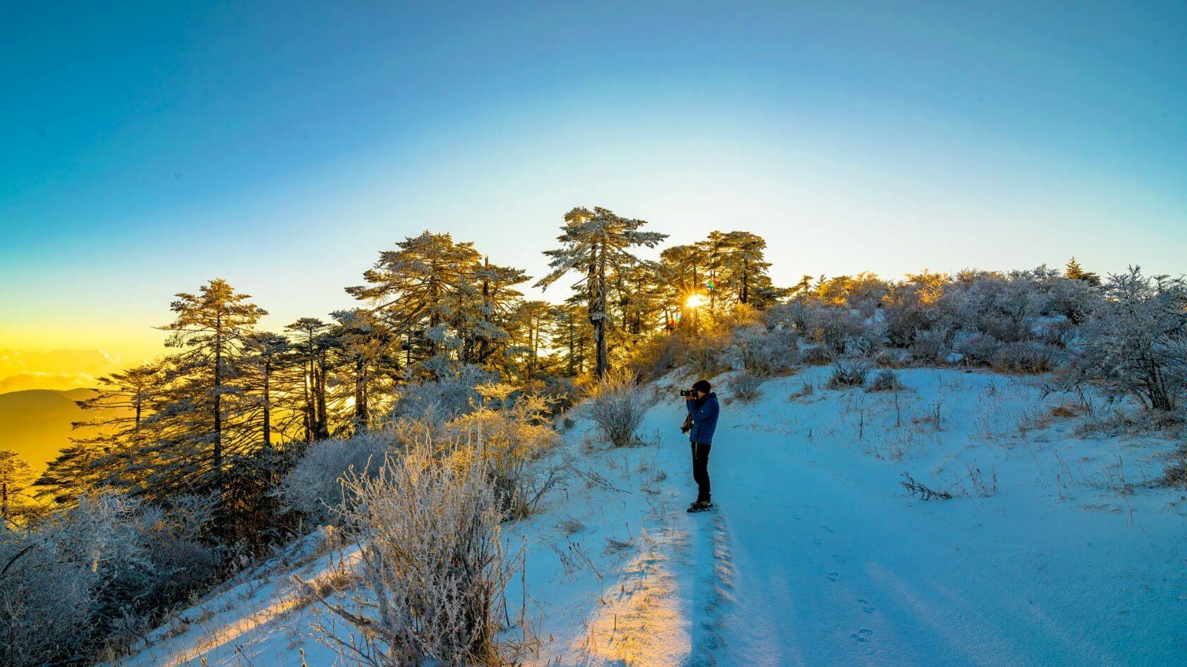 There is a man standing on top of a snowy hill wearing warm jacket, muffler and a hat tacking pictures of the distant view as sun is rising from his right covered with trees.